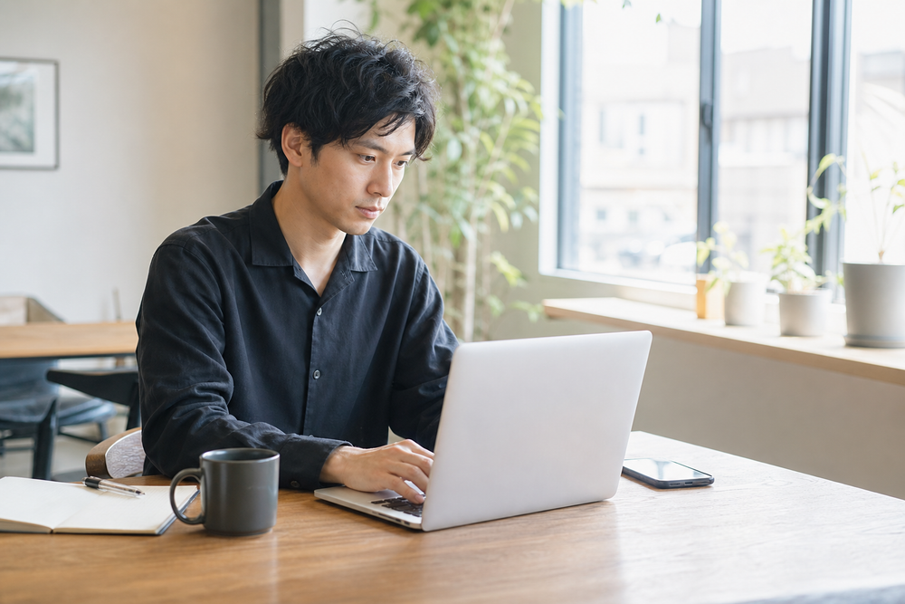 カフェや自宅の一室でノートパソコンに向かい、真剣な表情で作業をしているフリーランス風の個人事業主・クリエイターの写真風画像。自然光が差し込む明るい雰囲気、デスクにはノート、コーヒーカップ、スマートフォンが置かれている構図