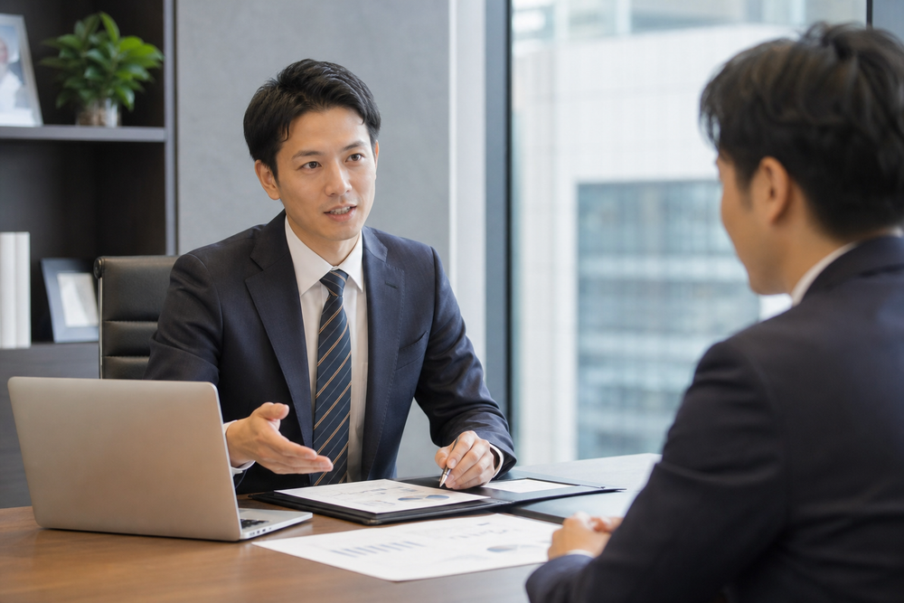 パソコンと書類資料を前に、専門家が顧客に対してコンサルティングや相談業務を行っているオフィスシーンの写真風イメージ、スーツ姿、落ち着いた雰囲気